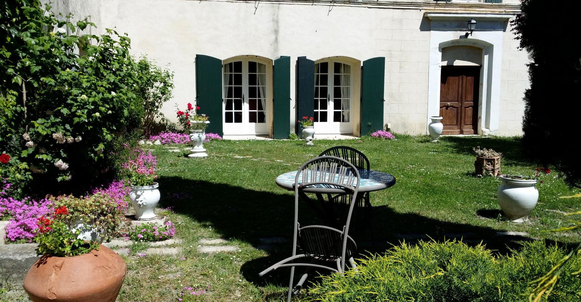 Jardin fleuri paisible au Mas Du Juge en Cammargue, terrasse ombragée avec table et chaises devant maison en pierre claire,